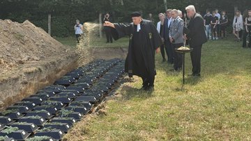 Drei lange Sargreihen in einer Grube: Davor steht ein Pfarrer und wirft Erde auf die Särge. Im Hintergrund stehen mehrere Menschen.