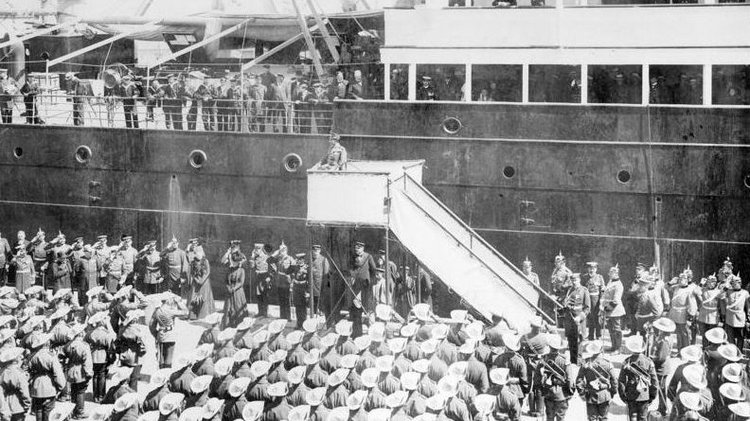 Auf einer Tribüne vor einem Schiff steht der Kaiser und spricht zu Soldaten und Matrosen, die auf und vor dem Schiff stehen.