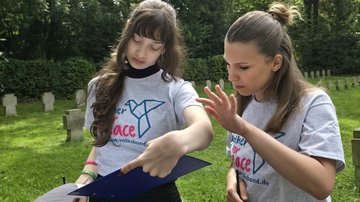 Zwei junge Frauen stehen auf einem Friedhof und besprechen einen Text