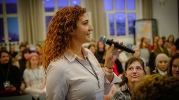 Junge Frau mit Mikrofon steht im Vordergrund. Im Hintergrund sitzen junge Menschen.