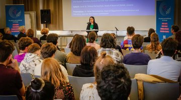 Ein Konferenzraum: Auf Stühlen sitzen junge Menschen, am Rednerpult steht eine Frau.