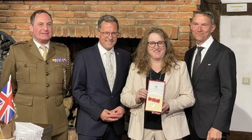 Drei Männer und eine Frau stehen vor einem alten Kamin. Die Frau zeigt eine Medaille in einer Kiste.