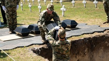 Eine Grabgrube: Ein Soldat übergibt einem anderen Soldaten einen Pappsarg.