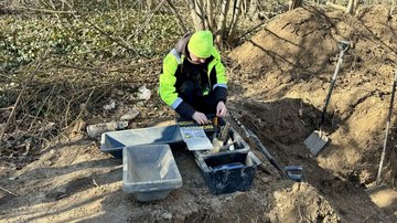 In einem Waldstück kniet ein Mann vor Kisten mit Werkzeugen.
