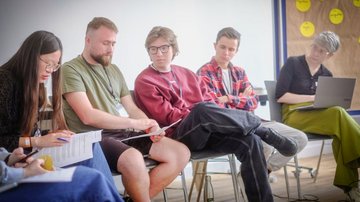 Vier junge Menschen sitzen auf Stühlen, rechts daneben sitzt eine Frau mit Laptop
