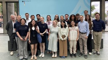 Eine Gruppe junger Menschen steht in zwei Reihen vor einer Schauwand des Außenministeriums.