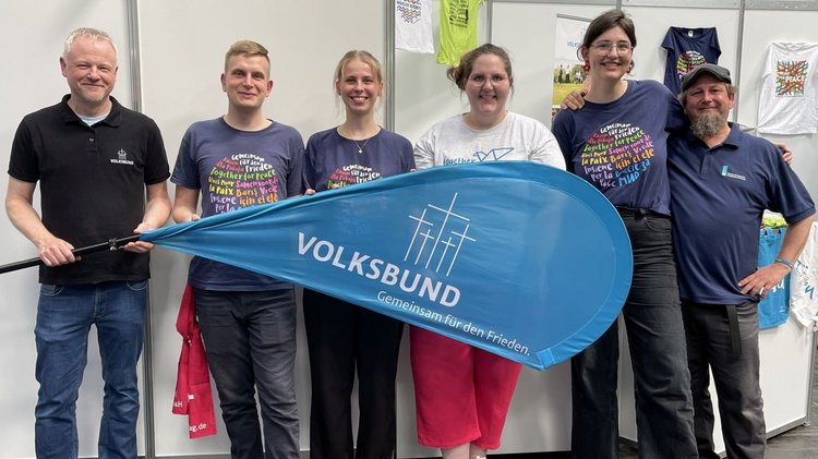 Drei Frauen und drei Männer unterschiedlichen Alters stehen vor einer Wand und halten eine Volksbund-Fahne