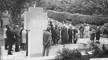 Ein terrassenförmiger Friedhof, im Vordergrund ein Hochkreuz aus Stein, im Halbkreis steht eine Gruppe zu einer Andacht