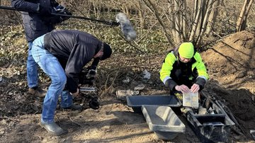 Ein Kameramann filmt einen Mann in Arbeitskleidung, der vor Kisten mit Werkzeug auf dem Waldboden kniet.