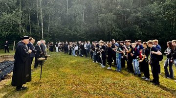 Am linken Bildrand: Särge in einer Grube, davor stehen Pfarrer und Männer in Anzügen, ihnen gegenüber steht eine große Gruppe Jugendlicher mit weißen Rosen