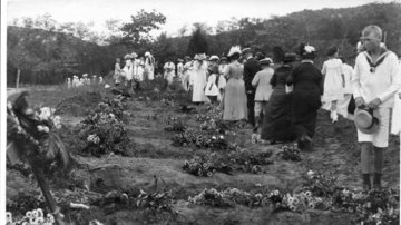 Blumen liegen auf Gräbern. Frauen und Kinder gehen über einen Friedhof. Ein Junge steht im Vordergrund an einem Grab.