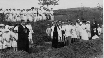 Zwei Pfarrer und eine Gruppe aus Frauen und Kindern stehen auf einem Friedhof.