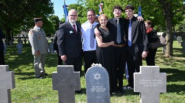 Kleine Gruppe auf einem Friedhof. Im Vordergrund: drei Grabkreuze und eine Grabstele.